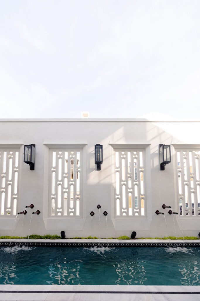 A row of water spouts pours into a narrow pool beneath a white wall with decorative precast panels and three black wall lanterns.