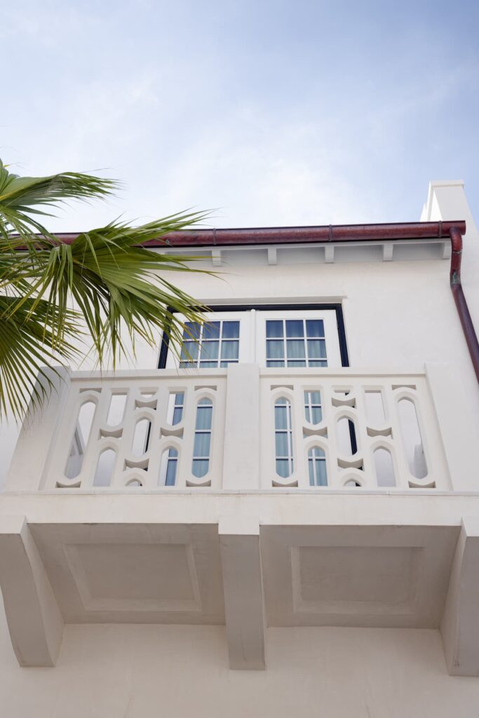 White balcony with decorative railing and double glass doors on an upper floor of a building, partially shaded by a palm tree, against a blue sky.