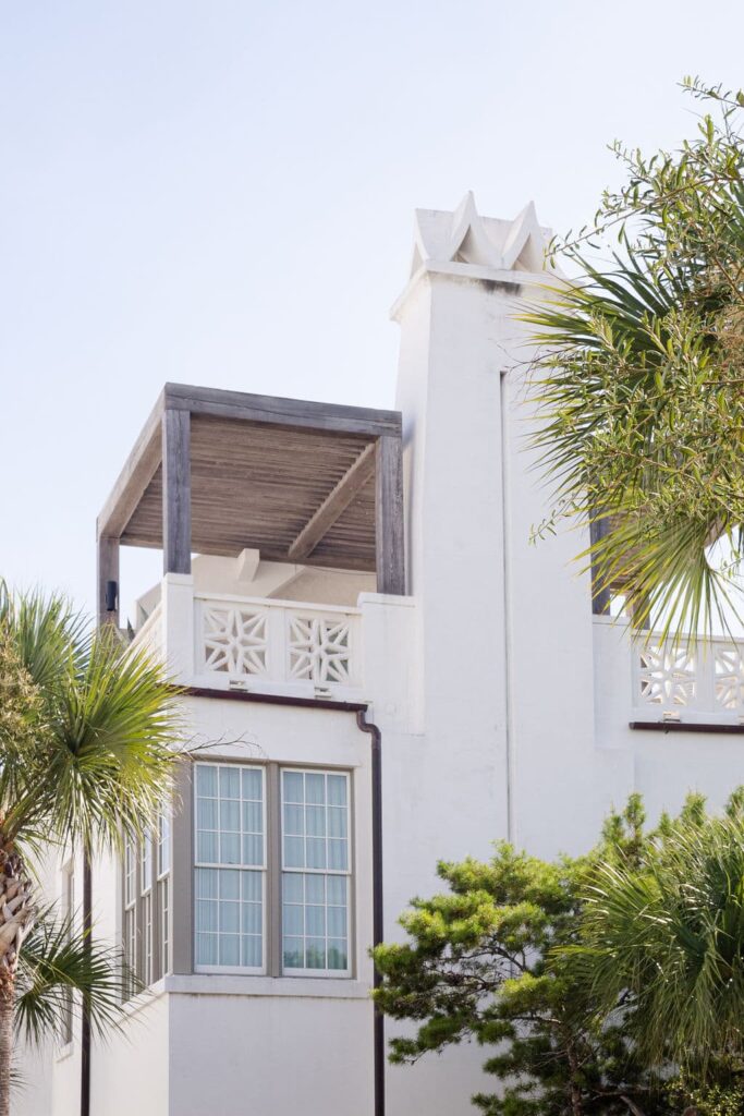 White stucco building with geometric architectural details, large windows, and a wooden balcony, framed by palm trees.