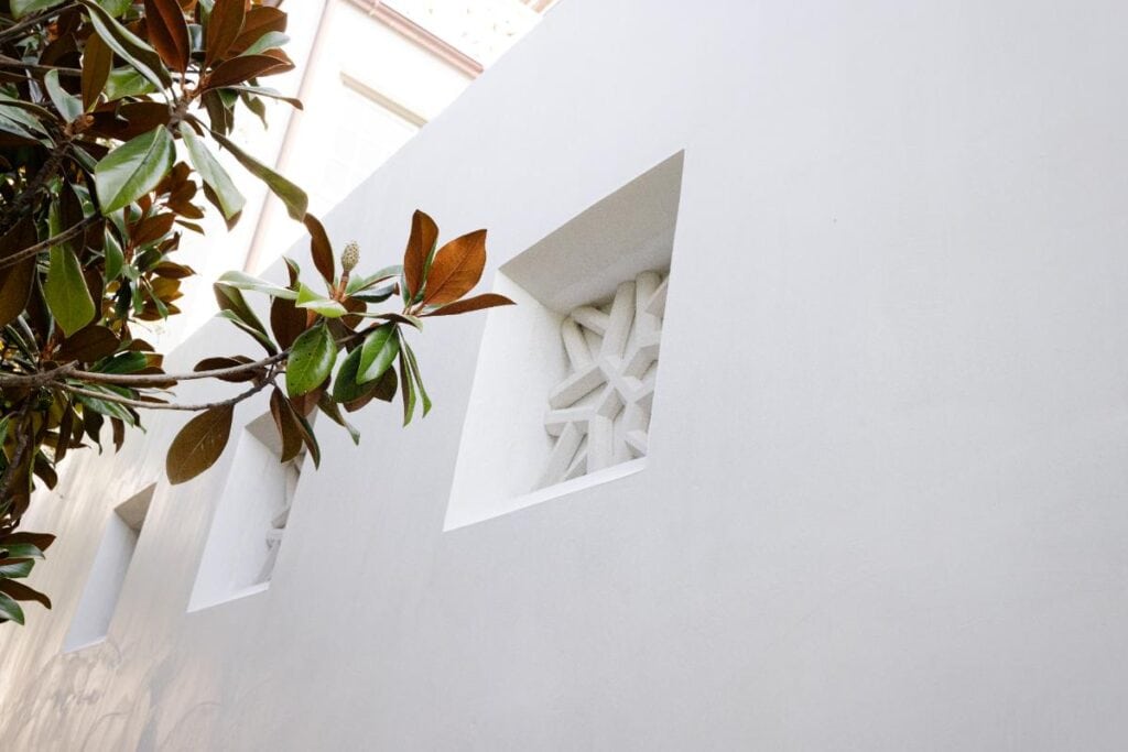 0S1A5461 A white wall with square precast vents is partially shaded by the branches and leaves of a nearby tree.