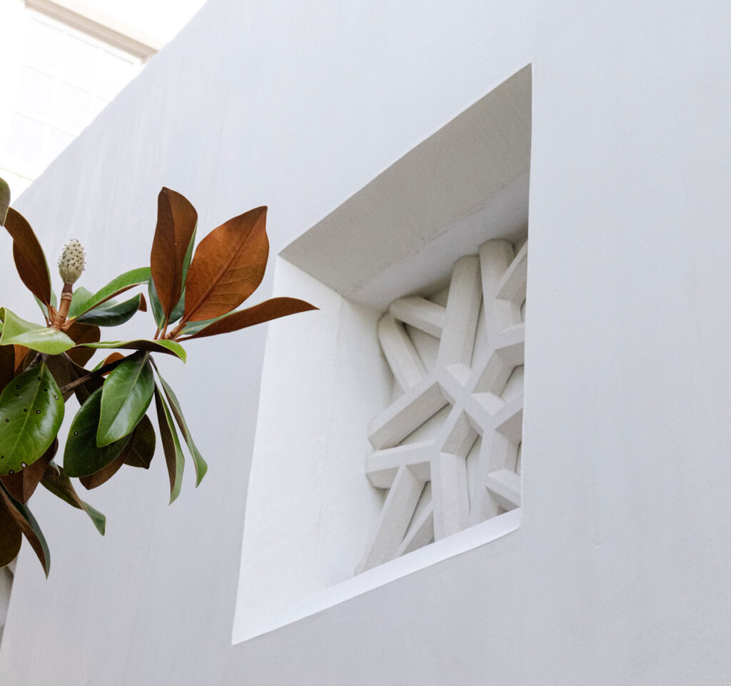 0S1A5462 A close-up of a white wall featuring a geometric decorative architectural precast feature recessed in a square opening, with green magnolia leaves and a cone extending from the left.