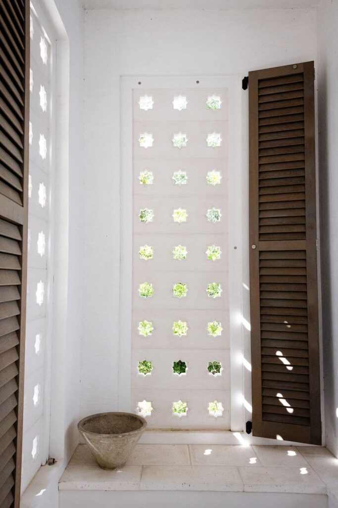 0S1A5518 (1) A white wall with architectural precast cutouts lets in light; wooden shutters are partially open on each side, and a ceramic bowl sits on a ledge below the window.