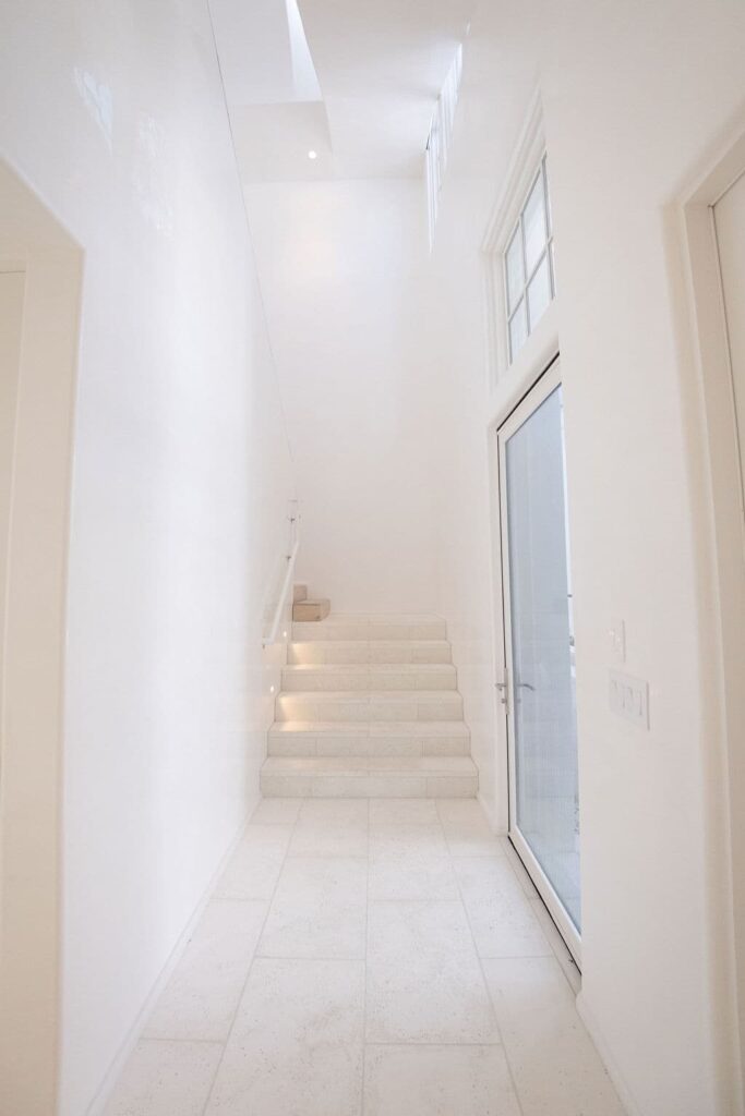 A white hallway with paver flooring, a staircase with white steps and railing, and a glass door on the right side.