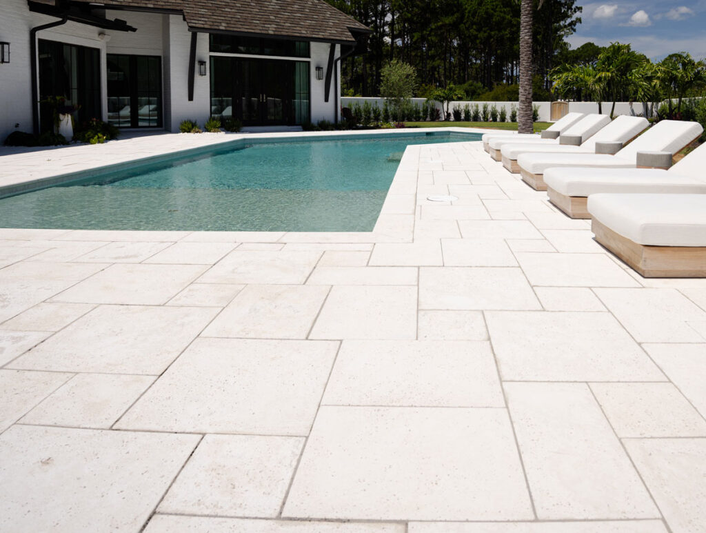 A modern outdoor swimming pool with white concrete paver decking and several white cushioned lounge chairs, next to a contemporary house.