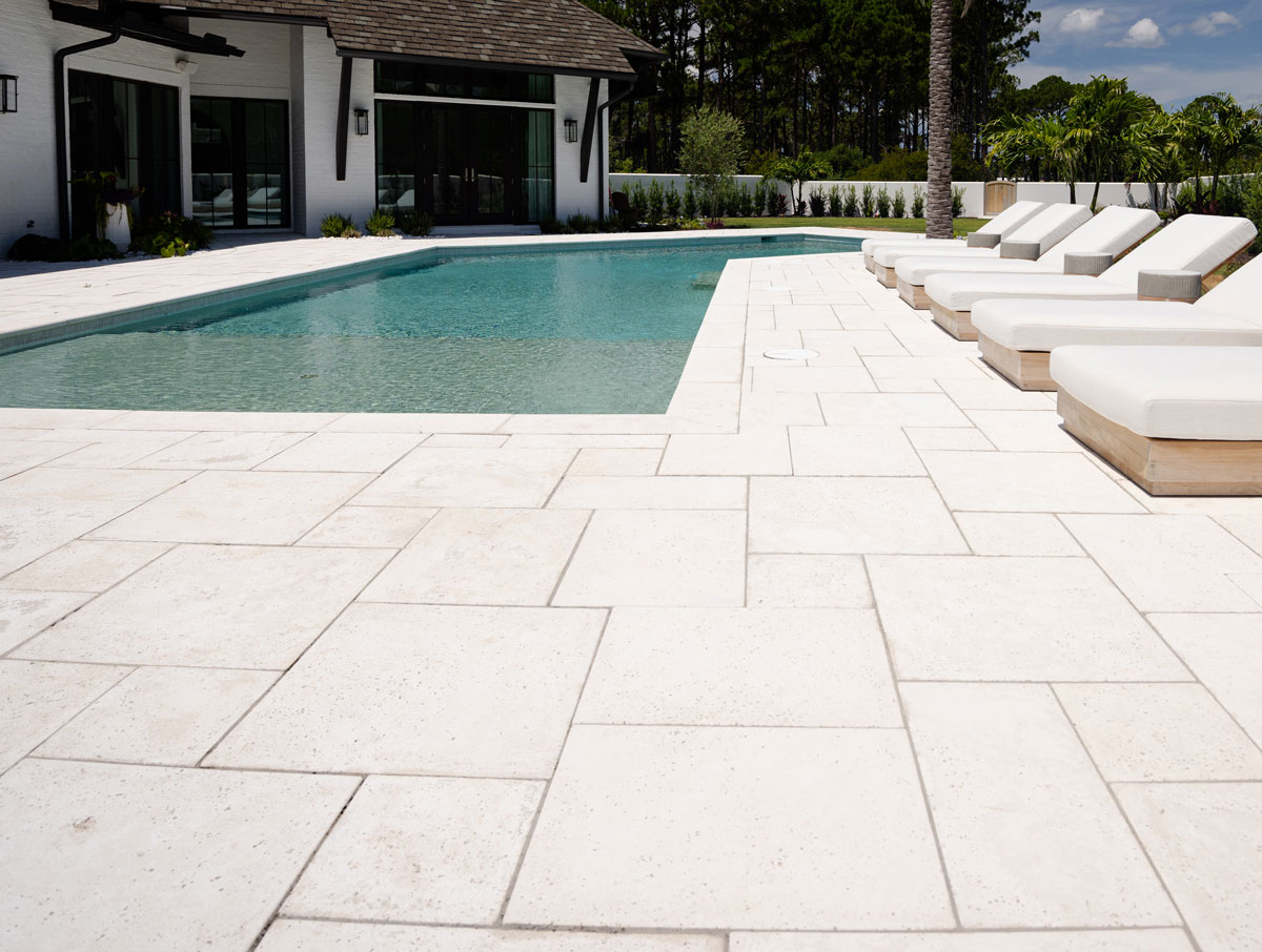 A modern outdoor swimming pool with white concrete paver decking and several white cushioned lounge chairs, next to a contemporary house.