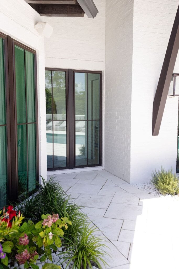 A white brick exterior corner of a house with tall black-trimmed windows, green plants, and a view of a pool through the glass doors.