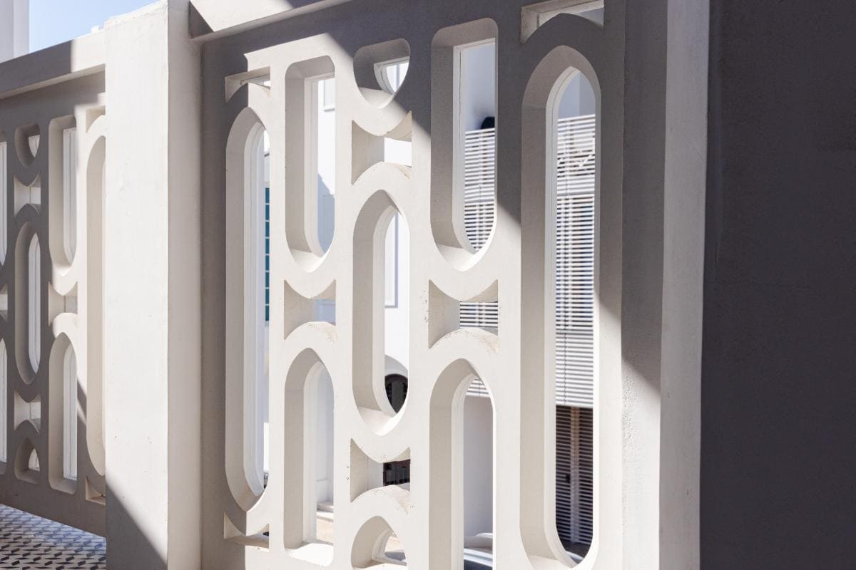 Balcony Railing 1 White concrete railing with geometric precast patterns, casting shadows in sunlight; architectural details and partial wall visible in the background.