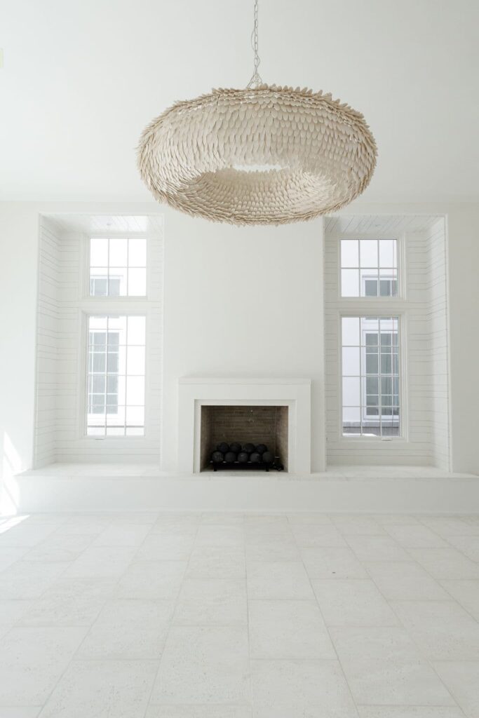 Minimalist white room with two tall windows, a simple fireplace, and a large woven pendant light hanging from the ceiling.