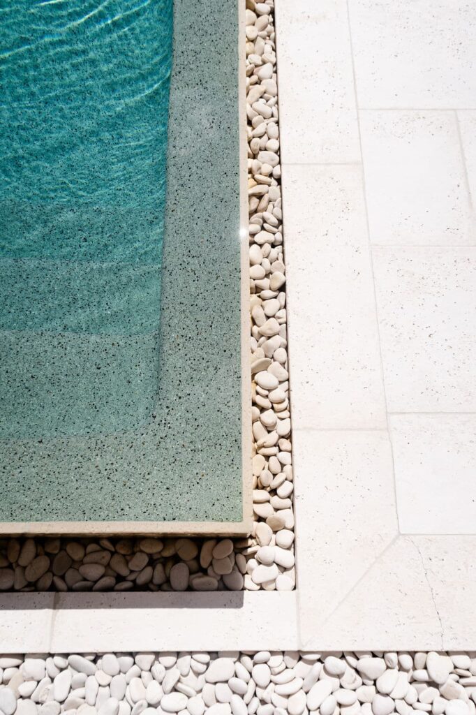 A close-up view of a swimming pool corner with turquoise water, beige concrete paver edge, white pebbles, and light-colored tiled flooring.
