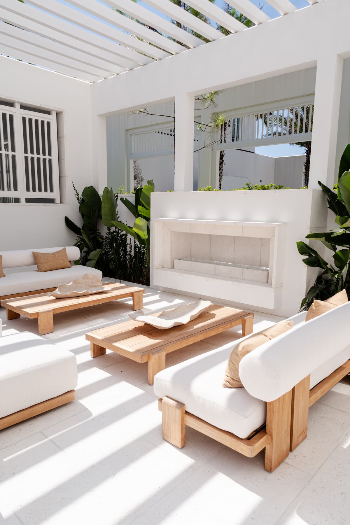 Modern outdoor patio with white cushioned wooden furniture, coffee tables, potted plants, and a fireplace under a slatted pergola. Natural light creates shadows on the floor.