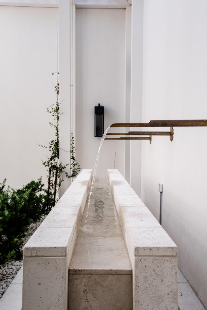 IMG_0684 A modern stone water trough with a brass spout extending from a white wall, water flowing into the trough, and greenery in the background.