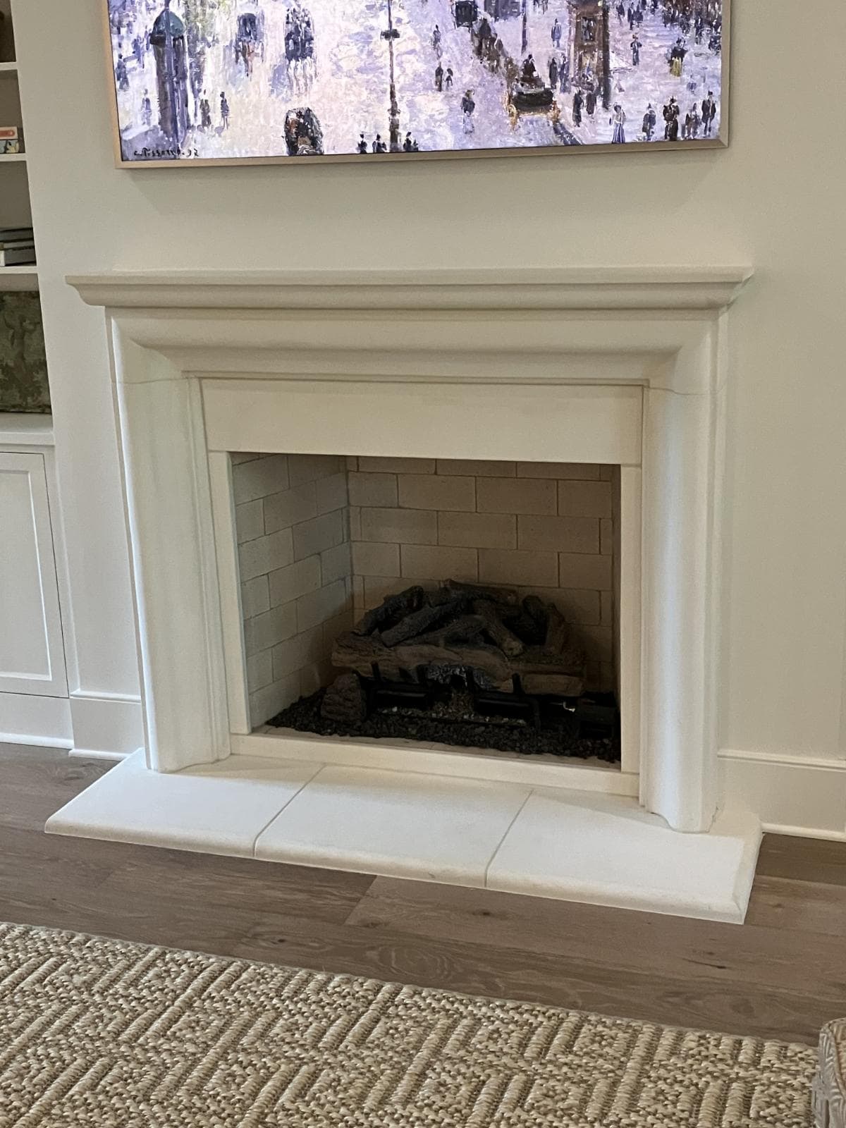 Interior Surround and Hearth 1 A concrete fireplace surround with a gas log set sits against a cream-colored wall beneath a painting. A textured beige rug and wooden floor are visible in the foreground.