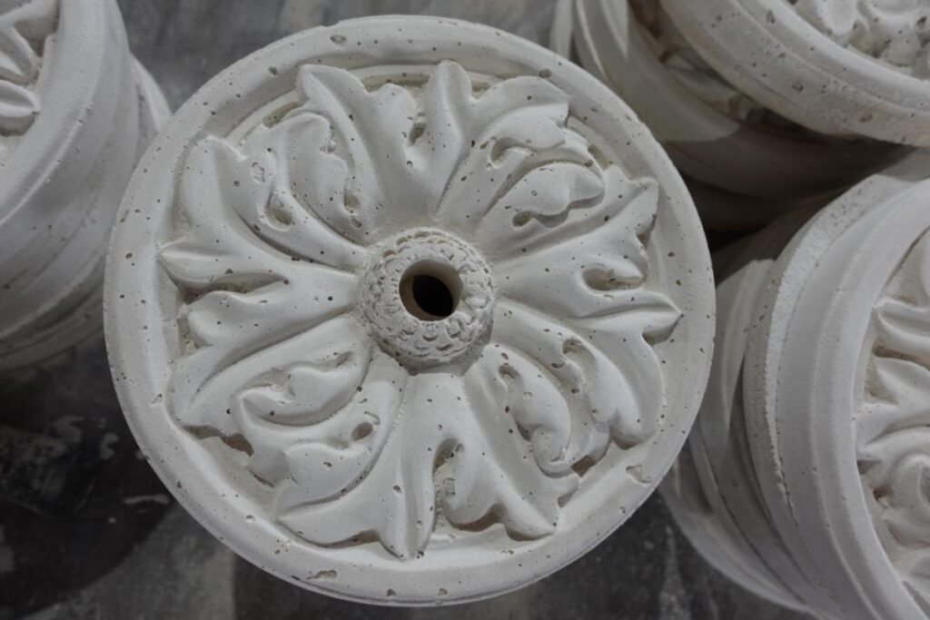 Rosettes Circular white concrete rosette with a central hole and raised, symmetrical leaf patterns, viewed from above. Several similar ornaments are partially visible around it.