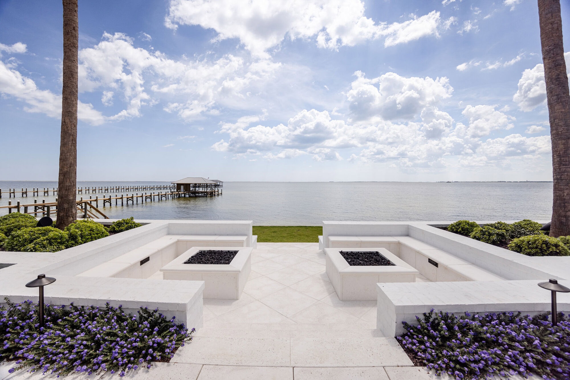 A waterfront patio with two white concrete fire pits, seating areas, and landscaped flowers overlooks a calm body of water and a wooden dock under a partly cloudy sky.