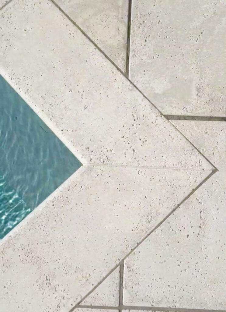 A geometric corner of a swimming pool shows textured paver tiles meeting at an angle above rippling blue water.