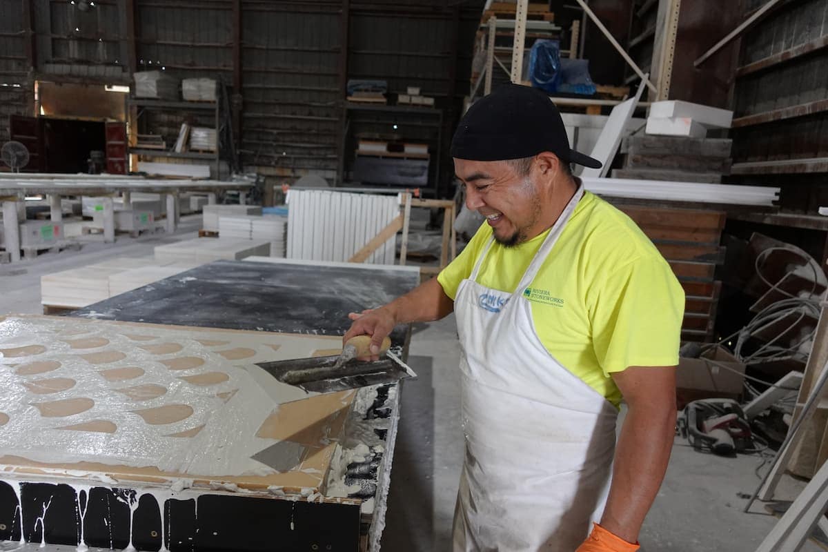 team member A man wearing an apron and gloves spreads concrete onto a mold using a trowel inside the Riveiera Stoneworks workshop.
