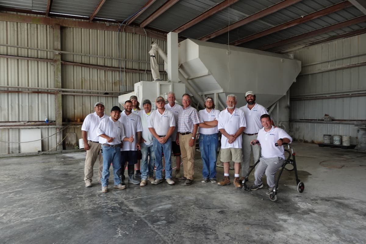 team The Riviera Stoneworks team wearing casual work clothes and white shirts stand in a warehouse in front of large industrial equipment.